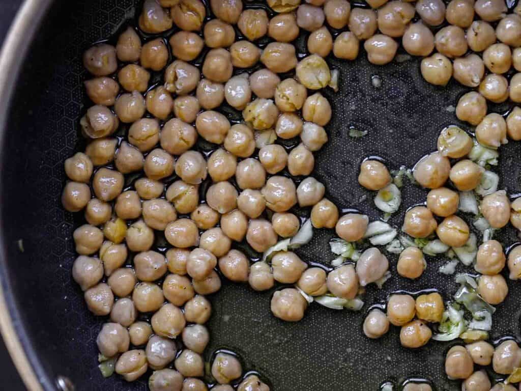 Chickpeas and chopped garlic cooking in a black pan with oil, a close-up view showing the chickpeas evenly distributed and glistening in the light.